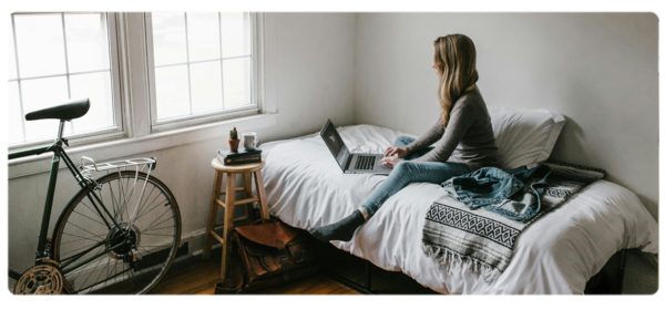 A student in a foreign country sits on a dorm bed and does homeowork on a laptop. In the corner, a bicycle is stored.